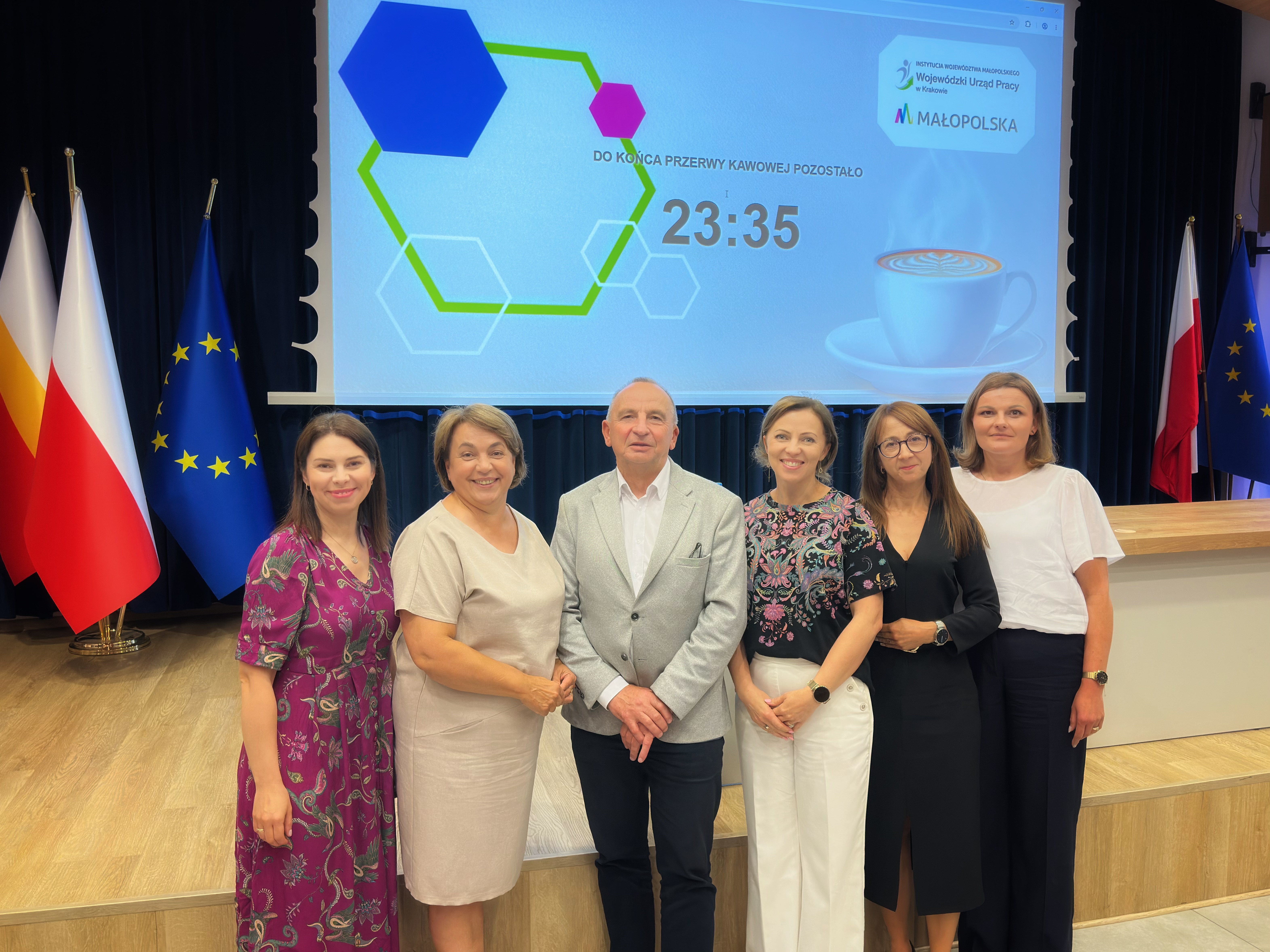Zdjęcie grupowe na scenie. Uczestnicy konferencji stoją na scenie Akademii Tarnowskiej, w tle widoczny ekran prezentacyjny oraz flagi Polski i Unii Europejskiej.